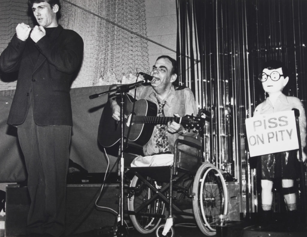 : Ian Stanton, a leading singer and songwriter in the Disability Arts Movement in the United Kingdom, performs onstage next to a BSL interpreter and “Chip the Crip,” a vintage Spastics Society collection box. Image courtesy of the National Disability Arts Collection and Archive (NDACA).