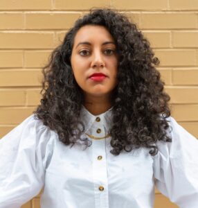 Diya Vij is a 30-something South Asian American woman with big black curly hair. She leans against a tan brick walls, staring directly into the camera, hands firmly on hip, with a red lip, gold jewelry and bossy white button down shirt.