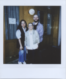 Photo ID: A polaroid of three people in front of a wood paneled storefront with glass windows and a blue curtain. Lu, an East Asian person stands in the center front, she is smiling and has her black hair pulled back and wears a white button up shirt, a tan cropped jacket, and large round glasses. Lee, a white man stands behind Lu smiling. He has a shaved head, brown beard, striped purple shirt, and silver hoop earring. Ixtzel, a Mexican woman, stands behind Lu, next to Lee. Ixtzel has loose black hair, a blue button-up, a black vest and white sneakers.