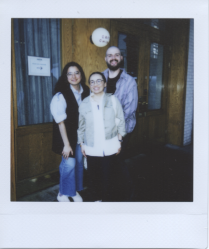 A polaroid of three people in front of a wood paneled storefront with glass windows and a blue curtain. Lu, an East Asian person stands in the center front, she is smiling and has her black hair pulled back and wears a white button up shirt, a tan cropped jacket, and large round glasses. Lee, a white man stands behind Lu smiling. He has a shaved head, brown beard, striped purple shirt, and silver hoop earring. Ixtzel, a Mexican woman, stands behind Lu, next to Lee. Ixtzel has loose black hair, a blue button-up, a black vest and white sneakers.